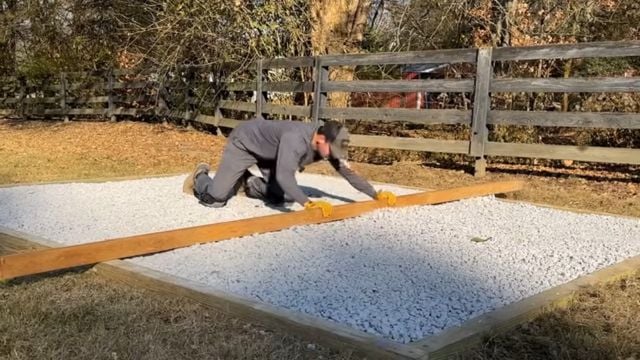 A man uses a piece of wood to level a gravel foundation.